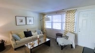 This two-floor space offers a comfortable and thoughtfully designed interior. The living area showcases a neutral color palette anchored by light beige walls and carpeted flooring, creating a warm and inviting atmosphere. A large window dressed with bold chevron-patterned curtains in gold and white serves as a focal point, allowing ample natural light to fill the room. Horizontal blinds provide adjustable privacy without sacrificing brightness.

The living space is furnished with a plush sofa adorned with decorative throw pillows in navy, teal, and green tones, complemented by a sleek mirrored coffee table that adds a touch of modern elegance. A cushioned accent chair with a draped throw blanket and a small dark wood side table provide additional seating options. A tall arc floor lamp offers functional yet stylish ambient lighting. Two framed artworks hang symmetrically on the wall, contributing to the curated aesthetic. A decorative vase with floral stems adds a natural element. The overall ambiance is cozy, contemporary, and well-balanced across both levels of the home.