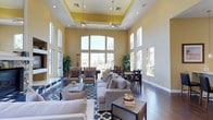 The clubhouse at 1390 Santa Alicia in Chula Vista is a thoughtfully designed communal space that blends elegance with functionality. The main gathering area immediately impresses with its dramatically soaring ceilings, accentuated by a raised tray ceiling detail finished in warm yellow tones. Dual ceiling fans ensure comfort throughout the space, while an expansive wall of arched and rectangular windows floods the interior with abundant natural light and offers views of the surrounding outdoors. The open-plan layout accommodates both relaxed lounging and social dining. The seating area features neutral-toned sofas and armchairs arranged around a central coffee table, anchored by a bold blue patterned area rug atop rich, dark hardwood flooring. Tasteful potted greenery adds a refreshing touch of nature to the interior. Adjacent to the lounge area, a dining space is outfitted with dark wood tables and cross-back chairs, ideal for community gatherings or casual meals. The warm yellow and white color palette throughout creates a welcoming and uplifting ambiance. Large windows lining the perimeter walls ensure the space remains bright and airy, making this clubhouse a versatile and inviting hub for residents.