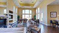 The clubhouse at 1390 Santa Alicia in Chula Vista is a thoughtfully designed communal space that blends elegance with functionality. The main gathering area immediately impresses with its dramatically soaring ceilings, accentuated by a raised tray ceiling detail finished in warm yellow tones. Dual ceiling fans ensure comfort throughout the space, while an expansive wall of arched and rectangular windows floods the interior with abundant natural light and offers views of the surrounding outdoors. The open-plan layout accommodates both relaxed lounging and social dining. The seating area features neutral-toned sofas and armchairs arranged around a central coffee table, anchored by a bold blue patterned area rug atop rich, dark hardwood flooring. Tasteful potted greenery adds a refreshing touch of nature to the interior. Adjacent to the lounge area, a dining space is outfitted with dark wood tables and cross-back chairs, ideal for community gatherings or casual meals. The warm yellow and white color palette throughout creates a welcoming and uplifting ambiance. Large windows lining the perimeter walls ensure the space remains bright and airy, making this clubhouse a versatile and inviting hub for residents.