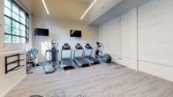 This fitness center presents a well-appointed, single-floor exercise space with a thoughtfully designed layout that accommodates both cardio and strength training needs. The space is divided into distinct functional zones, each equipped with professional-grade equipment.

The cardio area houses multiple treadmills and elliptical machines arranged in an organized row, with wall-mounted flat-screen televisions positioned for easy viewing during workouts. Linear LED lighting runs along the ceiling, providing bright, even illumination throughout. A secondary cardio zone features additional treadmills and elliptical machines, complemented by a wall-mounted television and access to natural light through windows.

The strength training area is outfitted with a dumbbell rack, a medicine ball storage tower, an adjustable workout bench, and a wall-mounted cable machine, offering a comprehensive range of resistance training options. Exposed concrete walls lend an industrial aesthetic to the space, contrasting with the clean white surfaces and durable patterned flooring. Black-framed glass doors and interior windows connect the fitness center to adjacent areas, maintaining an open, airy feel while defining the space clearly.