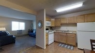This single-floor unit offers an efficient and functional layout, with the kitchen and living area connected in an open-concept arrangement. The kitchen is outfitted with light wood cabinetry providing ample storage, a white electric range with an overhead microwave, and a single-basin sink set into a continuous countertop. The tile flooring in the kitchen adds durability and ease of maintenance. A half-wall partition separates the kitchen from the adjacent living area, creating a sense of definition between the two spaces while maintaining an open feel. The living area features wall-to-wall carpeting and is brightened by a window that allows natural light to filter into the room. A wall-mounted decorative mirror adds a design accent near the partition. The textured ceilings are present throughout, and overhead fluorescent lighting in the kitchen ensures well-lit workspace. The overall ambiance is practical and straightforward, with a neutral color palette of beige and warm gray tones that lend a clean, modest aesthetic to the space.