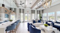 Located at 10196 SW Newberry Ave in Port Saint Lucie, FL, this elegantly designed single-floor community lounge offers a sophisticated coastal ambiance throughout its well-appointed layout. The space comprises a living room, kitchen, and hallway, all unified by light wood-look tile flooring and a soft, neutral color palette accented with deep navy blues.

The living room is the centerpiece of the space, boasting soaring ceilings adorned with exposed decorative wood beams and multiple ceiling fans. Expansive floor-to-ceiling windows dressed with floor-length drapes flood the interior with natural light while offering picturesque views of a resort-style pool and lush palm trees beyond. French doors provide direct outdoor access. The seating area features plush sofas, velvet swivel chairs, and a large-screen TV mounted on the wall, creating an inviting and functional gathering space. A built-in bar counter with upholstered stools lines one wall, ideal for casual seating.

The kitchen area is finished with crisp white cabinetry, open shelving, white quartz countertops, a stainless steel undermount sink, and white subway tile backsplash, with a window overlooking the landscaped grounds. A connecting hallway links the rooms seamlessly, completing this polished and welcoming community retreat.
