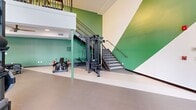 This impressive fitness facility, located at 5001 Palm Pointe Ct in Fort Myers, FL, is a testament to modern gym design. The space is spread across two floors, with the ground floor occupying 1810 square feet and the upper level covering 950 square feet. The gym's architecture is characterized by soaring ceilings and expansive windows that flood the space with natural light, creating an open and energizing atmosphere.

The facility boasts a wide array of cutting-edge fitness equipment, catering to various workout preferences. The main floor features a spacious open layout with dedicated areas for strength training, cardio exercises, and functional fitness. High-quality machines, free weights, and specialized equipment are strategically placed throughout the space. A notable design element is the incorporation of green accents on walls and support columns, adding a fresh and vibrant touch to the predominantly white interior.

The upper level, accessible via a modern staircase, overlooks the main floor, creating a dynamic, multi-level training environment. This design not only maximizes space utilization but also adds visual interest to the facility. The gym's layout promotes efficient traffic flow and allows for comfortable navigation between different workout zones.

Large garage-style doors on the ground floor suggest the possibility of an indoor-outdoor training experience, potentially opening up to exterior views or additional outdoor exercise areas. This thoughtful design enhances the overall functionality and appeal of this state-of-the-art fitness center.
