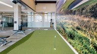 This dedicated indoor putting green offers a unique recreational amenity designed for golf enthusiasts. The space features a well-contoured artificial turf surface with multiple holes and flags, providing a realistic putting experience without leaving the building. A dramatic, large-scale photographic mural of a coastal golf course landscape spans an entire wall, creating an immersive and visually engaging atmosphere that enhances the feel of the green.

The architectural character of the space is defined by exposed brick walls, which lend a warm, industrial aesthetic that contrasts beautifully with the vibrant green turf. Natural light filters in through large windows with horizontal blinds, brightening the space throughout the day. The ceiling reveals industrial-style ductwork and exposed structural elements, consistent with the building's character.

Full-height glass walls and partitions separate the putting green from an adjacent fitness center, which is equipped with a comprehensive array of cardio machines, free weights, and weight benches. This transparent boundary maintains a visual connection between the two amenity spaces, creating an open and cohesive wellness environment. Modern pendant lighting supplements the natural light, ensuring the space is well-illuminated for use at any hour. Together, these features make this putting green a standout recreational amenity.