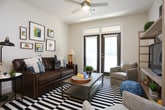 This single-floor residence offers a thoughtfully designed layout that balances comfort and modern style throughout. The living area serves as the heart of the home, featuring light-toned walls, durable hardwood-style flooring, and a bold black-and-white striped area rug. A set of dark-framed French doors with transom windows floods the space with natural light and provides outdoor access, while a gallery wall of framed abstract artwork adds a curated, artistic touch.

The kitchen is a standout feature, outfitted with grey shaker-style cabinetry, light granite countertops, a classic white subway tile backsplash, and stainless steel appliances including a range and microwave. A peninsula with an extended granite countertop and pendant lighting overhead creates a natural breakfast bar with seating for multiple guests, seamlessly connecting the kitchen to the main living space.

The bedrooms are carpeted and bright, with large windows that invite natural light and views of surrounding greenery. The primary bedroom includes a stylish sliding barn door that opens to a spacious walk-in closet with wire shelving and hanging rods. The bathroom is finished with granite-topped dark cabinetry, a large framed mirror, tile flooring, and a tub-shower combination. The walk-in closet provides generous storage, completing this well-rounded and functional living space.