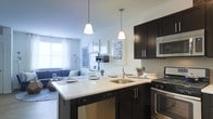 This single-floor space showcases an open-concept layout that integrates the kitchen and living area into one cohesive, functional environment. The kitchen is a standout feature, designed with rich dark cabinetry contrasted against a bright white countertop that extends into a peninsula-style breakfast bar, complete with pendant lighting overhead. Two sleek pendant lights hang at varying heights, providing focused task lighting while adding a contemporary design element. Stainless steel appliances, including a dishwasher, range, and built-in microwave, complement the dark cabinetry, while a classic white subway tile backsplash adds texture and a clean aesthetic. A stainless steel sink sits conveniently within the peninsula. The kitchen flows naturally into the living area, which benefits from natural light streaming through a large window dressed with white curtains. The living space features a neutral color palette with a blue sofa and curated wall art, creating a relaxed yet stylish atmosphere. The open layout enhances the sense of space, making the area feel both airy and connected throughout.