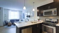 This single-floor space showcases an open-concept layout that integrates the kitchen and living area into one cohesive, functional environment. The kitchen is a standout feature, designed with rich dark cabinetry contrasted against a bright white countertop that extends into a peninsula-style breakfast bar, complete with pendant lighting overhead. Two sleek pendant lights hang at varying heights, providing focused task lighting while adding a contemporary design element. Stainless steel appliances, including a dishwasher, range, and built-in microwave, complement the dark cabinetry, while a classic white subway tile backsplash adds texture and a clean aesthetic. A stainless steel sink sits conveniently within the peninsula. The kitchen flows naturally into the living area, which benefits from natural light streaming through a large window dressed with white curtains. The living space features a neutral color palette with a blue sofa and curated wall art, creating a relaxed yet stylish atmosphere. The open layout enhances the sense of space, making the area feel both airy and connected throughout.