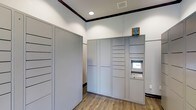 This single-floor package room is purpose-built to streamline the storage and retrieval of deliveries. The space is clean and well-organized, featuring white walls accented by dark crown molding trim that runs along the ceiling perimeter, giving the room a polished, finished appearance. Recessed lighting illuminates the interior with a bright, even glow.

The room is equipped with a bank of gray metal parcel lockers of varying sizes, accommodating packages of different dimensions. An automated locker kiosk with a touchscreen interface and keypad is integrated into the locker system, enabling secure, self-service package pickup. Additional gray storage lockers line the adjacent wall, providing further capacity for incoming deliveries.

The flooring is composed of wood-look vinyl planks in a warm, neutral tone, adding a touch of warmth to the otherwise utilitarian space. A wall-mounted light switch and a ventilation grille in the ceiling contribute to the room's functional design. Overall, this package room is thoughtfully designed to offer residents a secure, efficient, and easy-to-navigate solution for managing their deliveries.