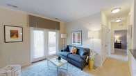 This single-floor space features a thoughtfully designed living area that balances comfort and style. The room is anchored by a blue-gray sofa adorned with colorful accent pillows, positioned atop a plush area rug that adds texture and warmth to the light-toned flooring. A sleek, minimalist coffee table sits in front of the sofa, providing both functionality and a clean aesthetic.

Natural light is a standout feature, streaming in through a set of French doors fitted with white horizontal blinds and a warm taupe Roman shade. The doors likely lead to an outdoor area, enhancing the sense of openness. Crown molding trims the ceiling throughout, adding an elegant architectural detail that elevates the overall finish of the space.

Lighting is well-considered, with both a table lamp and a tall floor lamp providing warm, ambient illumination. A framed piece of artwork on the wall adds visual interest, complementing the neutral color palette of soft whites and warm beiges. A louvered closet door is visible to the right, hinting at practical storage solutions. Overall, the space feels bright, airy, and inviting.