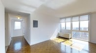 This single-floor space showcases a thoughtfully designed open layout that maximizes both light and livability. The main living area is anchored by rich herringbone parquet hardwood flooring that adds warmth and timeless character to the interior. Crisp white walls and white-trimmed ceilings keep the space feeling clean, airy, and versatile for a variety of furnishing styles.

A standout feature of the living area is the floor-to-ceiling window installation along one wall, flooding the space with abundant natural light and offering elevated city skyline views. A built-in radiator unit sits beneath the windows, providing heating while preserving the clean lines of the room. Sunlight casts bright, natural patterns across the parquet flooring, highlighting the quality of the wood.

An electrical panel is conveniently mounted on the interior wall, and a hallway connects the main living area to additional rooms beyond. The corridor, finished in the same neutral palette, leads further into the unit, suggesting additional functional spaces. Overall, the space presents a bright, well-maintained environment with classic architectural details and impressive urban views.