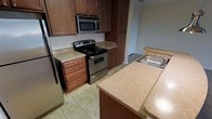 This single-floor space is designed with practicality and comfort in mind. The kitchen serves as a central highlight, showcasing rich, dark wood cabinetry that extends from base to upper levels, providing generous storage throughout. The cabinetry is complemented by light-toned laminate countertops that offer ample workspace for meal preparation, including an extended peninsula-style counter with a stainless steel sink. Stainless steel appliances, including an electric range with a built-in microwave above, add a modern touch and are thoughtfully integrated into the layout. Light-colored tile flooring runs throughout the kitchen, contributing to a clean and cohesive aesthetic. The layout is efficiently organized, allowing for easy movement and access to all areas. A doorway visible from the kitchen suggests a smooth, open flow into adjacent spaces, reinforcing the single-floor design's connectivity. Overall, the space conveys a straightforward, functional character with warm tones from the cabinetry balanced by lighter surfaces, making it a practical and inviting environment suited for day-to-day use.