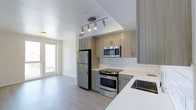 This single-story, two-bedroom, one-bathroom residence features a thoughtfully designed open layout that maximizes functionality and flow. The kitchen serves as a central highlight of the space, outfitted with stainless steel appliances including a refrigerator, range, and over-the-range microwave. Light wood-toned cabinetry lines the walls and provides ample storage, while a white subway tile backsplash adds a clean, contemporary contrast. Light-colored quartz countertops and a stainless steel sink complete the kitchen's polished look. Track lighting overhead ensures the workspace is well-lit. Light wood-style flooring runs throughout, lending a cohesive and modern aesthetic to the interior. A glass-paneled door at the far end of the space allows natural light to filter into the living area, creating a bright and airy atmosphere. The neutral color palette of white walls and soft, natural tones throughout contributes to an open, welcoming ambiance. The overall design reflects a modern sensibility with practical finishes suited for comfortable everyday living.