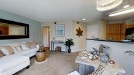 This 2-bedroom, 2-bathroom residence is designed with a coastal aesthetic that creates a warm and inviting atmosphere throughout. The open-concept living and dining area serves as the heart of the home, featuring plush carpeting, neutral wall tones with soft sage green accents, and recessed lighting that keeps the space bright and welcoming. Decorative touches such as nautical-themed décor, driftwood accents, and a framed ocean-view artwork reinforce the relaxed, beachy ambiance. The dining area flows naturally into the kitchen, which is elevated by dark countertops and a convenient breakfast bar with wicker seating, adding both function and character. A console table with decorative objects and lush indoor greenery adds depth and charm to the living area. The layout is efficient and well-organized, with clear sightlines connecting the main living spaces. The single-floor plan ensures easy accessibility throughout. Recessed and pendant lighting provide layered illumination, enhancing the overall warmth of the interiors. This residence is a comfortable and stylish home that balances casual coastal living with practical, everyday functionality.