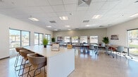 Located at 6775 N 67th Ave in Glendale, Arizona, this single-floor commercial space offers a bright, open, and functional layout well-suited for community gatherings, training sessions, or collaborative activities. The interior is defined by gleaming polished terrazzo-style concrete floors that reflect natural light throughout the room, contributing to an airy and welcoming atmosphere.

Upon entry, visitors are greeted by a clean, white reception desk accented by a welcoming sign, with the Arizona state flag and an American flag flanking the space near the entrance. Large glass entry doors allow views to an outdoor area featuring a playground and landscaping. The open main area is filled with modular tables and chairs arranged in flexible configurations, making the space highly adaptable for various uses.

Natural light pours in through multiple large windows lining the perimeter walls, offering views of the surrounding neighborhood. A drop-tile ceiling with recessed fluorescent lighting ensures consistent illumination throughout. A bold black-and-white geometric tile accent wall serves as a striking focal point near a kitchenette area stocked with countertop appliances. Potted tropical plants are strategically placed to add warmth and greenery to the otherwise minimalist interior. The overall design is clean, modern, and purposefully functional.