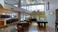This contemporary amenity lounge is defined by its striking double-height ceilings and expansive floor-to-ceiling windows that flood the space with natural light while offering open views of the surrounding outdoor greenery and adjacent buildings. The architectural design emphasizes openness and volume, with a glass-paneled upper mezzanine level adding a sense of grandeur to the interior.

The main floor is thoughtfully arranged into distinct activity zones. Dining and casual seating areas feature sleek, dark-framed tables paired with upholstered chairs in warm neutral tones, ideal for socializing or working. A bar-height counter with tall stools lines one side of the space, providing an additional gathering spot. Toward the far end of the room, a full-sized billiards table anchors a recreation zone, complemented by lounge seating arranged on a patterned area rug nearby.

Overhead, a series of modern cylindrical pendant and flush-mount light fixtures are evenly distributed across the ceiling, providing consistent, warm illumination throughout. The polished hardwood flooring ties the entire space together, reflecting the abundant natural light and enhancing the airy ambiance. Large sliding or fixed glass panels along the perimeter connect the interior visually to the outdoor landscaped areas, creating a seamless indoor-outdoor feel.