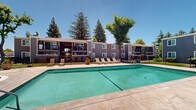 Located at 5109 Andrea Blvd in Sacramento, California, this outdoor amenity area serves as a central gathering space within a multi-building residential community. The centerpiece is a generously sized swimming pool with clear turquoise water, framed by a wide concrete pool deck that provides ample room for lounging and socializing. A separate circular hot tub/spa is positioned adjacent to the main pool, offering a relaxing retreat alongside additional seating. The pool area is enclosed by a sleek wrought iron fence with a gated entry, ensuring a secure and private environment for residents. Numerous lounge chairs and patio seating are arranged around the perimeter, ideal for enjoying Sacramento's sunny climate. Mature trees — including tall pines and deciduous varieties — surround the space, providing natural shade and a lush, park-like backdrop. Decorative planters with flowering plants and greenery add visual warmth to the patio. The multi-story residential buildings in gray and purple tones border the courtyard, with balconies overlooking the pool area. The open-sky setting allows for abundant natural light, making this an inviting outdoor destination throughout the day.