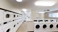 This communal laundry facility is designed for efficiency and ease of use, offering a generous array of machines to accommodate high-demand usage. Along one wall, a row of stacked dryer units maximizes vertical space, providing substantial drying capacity without compromising floor area. Opposite these, a lineup of front-loading washing machines offers ample washing capacity. A centrally positioned white countertop runs through the space, serving as a convenient folding and sorting surface. The room is finished in neutral tones throughout, with light-colored walls and flooring contributing to a clean, utilitarian aesthetic. Bright fluorescent ceiling fixtures ensure the entire space is well-lit, enhancing visibility and practicality. A structural column adds architectural definition to the room without interrupting the functional layout. A door at the far end provides access to an adjacent area, suggesting the facility is part of a larger building. Overall, the space is organized, functional, and designed to serve the laundry needs of multiple users efficiently.