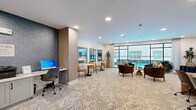 Located at 1888 Brittmoore Rd in Houston, Texas, this well-appointed community amenity space offers a thoughtfully designed single-floor layout that seamlessly blends work and leisure. The space is finished with a neutral herringbone-patterned carpet that runs throughout, complemented by crisp white ceilings with recessed lighting that keeps the entire area bright and welcoming.

The lounge area is anchored by large floor-to-ceiling windows with sleek black frames, offering direct views of the outdoor pool and filling the space with abundant natural light. Comfortable brown leather armchairs and cozy seating clusters are arranged to encourage relaxed conversation, while marble-topped bistro tables paired with amber leather chairs provide intimate spots for casual meetings or dining.

Bold, gold-framed abstract artwork adorns the walls, adding vibrant pops of color against the clean white and textured blue accent walls. A dedicated business center features dual computer workstations set against a striking deep-blue textured feature wall, complete with printing capabilities for residents' convenience.

A conference table with ample seating and a wall-mounted display screen creates a functional collaborative workspace near the glass entry doors. A package locker system is also integrated near the entrance, adding a practical touch to this polished, amenity-rich space.