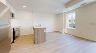Located at 97 Milford Ave in Waterloo, Ontario, this contemporary two-storey home offers a well-designed layout spread across a generously sized main floor and a more compact lower level. Throughout the interior, light-toned hardwood-style flooring creates a cohesive and bright aesthetic, complemented by crisp white walls and clean white trim.

The main floor is the heart of the home, featuring an open-concept living and kitchen area anchored by a freestanding island with a wood-grain finish and integrated sink — an ideal setup for both everyday living and entertaining. Large windows flood the space with natural light and offer views of the surrounding neighbourhood. A bright bedroom or flex room on this level includes sliding glass doors that open directly onto a private balcony enclosed by modern glass railings, providing an airy outdoor retreat.

Additional rooms on the main floor include well-proportioned bedrooms with ample natural light from dedicated windows. A hallway bathroom features a wood-grain vanity with a white countertop, maintaining the warm, modern tone found throughout.

The lower level offers versatile open space with recessed lighting, a staircase with dark-toned treads and a sleek handrail, and a dedicated utility room housing a stacked washer/dryer and water softener system. This level provides excellent potential for a recreation room, home office, or additional living space.
