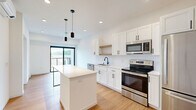 Located at 100 N Farwell St in Eau Claire, Wisconsin, this thoughtfully designed one-bedroom, one-bathroom unit offers a clean, modern aesthetic throughout its efficiently arranged floor plan. The open-concept kitchen and living area serves as the heart of the unit, featuring white shaker cabinetry, a classic subway tile backsplash, a natural wood open shelf, and a full suite of stainless steel appliances including a range, microwave, dishwasher, and refrigerator. A center island with a white quartz-style countertop provides additional prep space and casual seating, accented by sleek pendant lighting above. Warm bamboo hardwood flooring runs throughout the main living area, complementing the bright, white walls and recessed lighting. A sliding glass door opens to a private balcony, inviting natural light and outdoor views into the living space. The bedroom is a serene, minimalist retreat with large floor-to-ceiling windows framing outdoor views and a modern barn-style sliding door leading to a closet. The bathroom features a warm wood vanity with white countertop, matte black fixtures, a large mirror, and neutral tile flooring, offering a spa-like, functional finish.