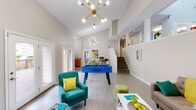 This energetic and thoughtfully designed space makes a strong impression with its soaring vaulted ceilings and an open-concept layout that seamlessly connects the living, dining, and kitchen areas. The main living room is anchored by a striking stone fireplace topped with a rustic wooden mantel and a wall-mounted flat-screen TV, flanked by teal accent chairs and a tufted sofa adorned with vibrant throw pillows. A bold Sputnik-style gold chandelier adds a mid-century modern flair, illuminating the light gray hardwood-style flooring throughout.

A foosball table positioned centrally in the open gathering area adds a playful, recreational dimension to the space. The dining area features a warm wood table set for multiple guests, accented with yellow floral arrangements and colorful dishware, creating a welcoming atmosphere for shared meals.

The kitchen is well-appointed with warm honey-toned cabinetry, granite-style countertops, stainless steel appliances including a refrigerator and electric range, and a stainless steel sink beneath a bright window. Interior pass-through windows connect the kitchen and living zones visually, enhancing the open feel.

A staircase leads to an upper level, visible through clerestory-style windows. Additional amenities visible from the layout include a fitness area. The overall ambiance is lively, modern, and designed for communal use.