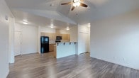 This single-floor space showcases a clean, modern design centered around an open-concept floor plan that seamlessly connects the living area to the kitchen. The kitchen is equipped with light wood cabinetry, black appliances including a full-size refrigerator and microwave, and a peninsula counter with bar-top seating — ideal for casual dining or entertaining. The peninsula serves as a natural divider between the kitchen and the main living area, maintaining an airy, connected feel throughout the space. Durable hardwood-style flooring runs throughout, adding warmth and a contemporary touch underfoot. The living area features a vaulted ceiling with a ceiling fan and integrated lighting, enhancing the sense of height and openness. Recessed lighting throughout ensures the space is well-lit. Crisp white walls keep the interior feeling bright and clean, providing a neutral backdrop that complements a variety of furnishing styles. A closet with double doors offers convenient built-in storage. The overall ambiance is fresh, functional, and modern, making this a well-appointed space suited for comfortable everyday living.