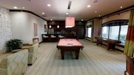 This single-floor recreational lounge is thoughtfully designed to serve as a premier entertainment destination. The centerpiece of the space is a full-size billiards table with a warm salmon-toned felt surface, positioned beneath a bold square pendant light that anchors the room visually. Rich dark wood tones carry throughout the space, from the cabinetry to the pool table's sturdy carved legs, lending the lounge a sophisticated yet inviting character.

Along the back wall, a wet bar area features dark wood cabinetry with a mosaic tile backsplash, a stainless steel refrigerator, and a sleek countertop lined with white bar stools — ideal for hosting guests. Additional pendant lighting above the bar adds warmth and depth to this section of the room.

Natural light filters in through draped windows along one wall, softening the interior atmosphere while complementing the light-toned, wood-look flooring that runs throughout the space. A shuffleboard table is positioned along the far wall, offering yet another avenue for recreation.

Recessed ceiling lighting provides even illumination across the room, while decorative artwork on the walls adds personality and color. A comfortable seating area with upholstered furniture completes the lounge, making this space as relaxing as it is entertaining.