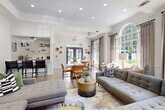 This elegantly designed single-floor community space blends modern and transitional aesthetics to create an inviting, well-appointed environment. The entry foyer features a sleek white quartz reception desk, an artistic sculptural pendant light, exposed whitewashed brick accent wall, and floor-to-ceiling glass doors that draw in views of the surrounding greenery.

The expansive main lounge is defined by tray ceilings, recessed lighting, and shiplap-clad walls. A gray brick fireplace anchors one end of the room, topped by a wall-mounted television, while generous tufted gray sectionals and gold drum accent tables furnish the space. Large multi-pane windows and an arched transom window flood the room with natural light and offer leafy outdoor views.

Adjacent to the lounge, a casual dining area features a round white table with warm-toned chairs, creating an intimate yet open gathering space. The kitchenette is thoughtfully designed with slate-gray shaker cabinetry, white quartz countertops, an undermount sink, beverage refrigerator, coffee station, and open shelving adorned with curated décor and plants.

Throughout, light wood-look flooring, neutral drapery, gold metallic wall accents, and vibrant original artwork contribute to a cohesive, upscale ambiance suited for community gatherings or professional interactions.