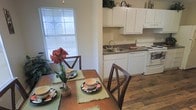 This single-floor space offers a seamlessly connected dining and kitchen layout, creating an open and functional living environment. The kitchen is equipped with white cabinetry that extends to the upper walls, providing ample storage space, including cabinets positioned above the appliances for additional organization. The countertops feature a dark laminate surface, complemented by a stainless steel sink and white appliances, including an electric range and dishwasher. Upper cabinets are topped with decorative greenery, adding a touch of warmth to the space. The kitchen flows naturally into the dining area, which is anchored by rich, hardwood-style plank flooring that adds character and warmth throughout. The dining space accommodates a wooden table set with place settings and is adorned with a bold red floral centerpiece, contributing a welcoming ambiance. A large window with horizontal blinds allows natural light to fill the area, enhancing the bright and airy feel of the space. The neutral wall tones throughout provide a clean backdrop that ties the kitchen and dining areas together cohesively.