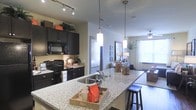 This single-floor space showcases a thoughtfully designed open-concept layout where the kitchen and living area connect seamlessly. The kitchen is a standout feature, equipped with dark espresso cabinetry that extends to the upper walls, providing generous storage. A large granite-topped island anchors the space, complete with an undermount sink and bar seating, making it ideal for both meal preparation and casual dining. Pendant lighting with sleek cylindrical shades hangs above the island, adding a modern touch, while a track lighting system illuminates the kitchen workspace. Black appliances, including a range and built-in microwave, complement the bold cabinetry. The countertops carry the same speckled granite finish throughout, creating visual continuity. The open layout transitions naturally into the living area, which benefits from abundant natural light streaming through large windows dressed with light curtains. A ceiling fan keeps the living space comfortable. The overall ambiance balances contemporary design with functional living, highlighted by the contrast between dark cabinetry, light granite surfaces, and neutral-toned walls throughout.