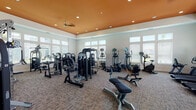 Located at 2301 Academy Circle West in Kissimmee, Florida, this fitness center is designed to support a comprehensive workout experience within a single-story facility. The main exercise area is defined by its distinctive tray ceiling, finished in a bold orange accent and equipped with recessed lighting and a ceiling fan for enhanced comfort. Neutral-toned carpet flooring spans the primary workout space, which is lined with large windows fitted with adjustable blinds, inviting generous natural light while maintaining views of the surrounding outdoor environment.

The strength training area is well-stocked with a variety of cable and selectorized resistance machines, adjustable benches, and multi-function weight stations, providing options for full-body training. The cardio section features a row of treadmills positioned directly in front of expansive windows, alongside ellipticals, stationary bikes, a rowing machine, and a spin cycle, all equipped with individual display monitors. A separate studio area connects via a hallway and offers hardwood-style flooring, additional windows, and open space suitable for stretching, functional training, or group exercise with stability balls and accessories. Accent artwork and a bench in the entryway corridor contribute to a polished, welcoming atmosphere throughout the facility.