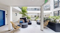 Located on the second floor of a modern residential building in Los Angeles, this thoughtfully designed outdoor amenity space offers a refined and inviting environment centered around an open-air courtyard. The courtyard is framed by the building's upper balconies and clean white facades, creating a semi-enclosed atmosphere that balances privacy with openness to the California sky. Lush tropical and subtropical plantings, including large-leafed monstera, ornamental grasses, and mature trees set within elevated planters, bring a vibrant, garden-like quality to the space. The lounge area is furnished with woven rattan accent chairs, low-profile modular sofas, and round terrazzo-style coffee tables, offering a comfortable and stylish setting for relaxation or socializing. Light-toned tile flooring throughout reflects natural light and reinforces the space's bright, airy character. Structural columns provide a covered section of the lounge, offering shade while maintaining visual connection to the open courtyard. An additional exterior terrace extends from the main space, furnished with sleek dining tables and chairs, and framed by a textured white brick wall with views of surrounding palm trees and the Los Angeles neighborhood beyond. Elevator access is visible from within the courtyard, ensuring convenient connectivity to the rest of the building.