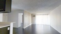 This single-floor space offers a clean, functional layout with a comfortable, no-frills aesthetic. The open living area is a standout feature, showcasing rich, dark hardwood flooring that contrasts cleanly with the neutral beige walls and classic textured ceiling. Large sliding glass doors dressed with sheer white curtains line one wall, inviting abundant natural light and creating a bright, airy atmosphere throughout the main living space. A wall-mounted air conditioning unit ensures year-round climate comfort. A wide doorway leads to an adjoining room, hinting at a practical, open-flow floor plan that connects the living area seamlessly to the rest of the space. The clean white baseboards and trim provide a crisp finishing touch throughout. The overall ambiance is understated and move-in ready, offering a neutral canvas well-suited for a variety of interior styles. The single-floor layout provides convenient, step-free access throughout, making the space highly accessible and easy to navigate.