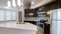 This single-floor space showcases a thoughtfully designed interior that blends contemporary aesthetics with practical functionality. The kitchen serves as a standout centerpiece, featuring rich dark cabinetry that extends to the upper walls, offering generous storage throughout. Complementing the cabinetry is a sweeping granite island countertop in a light speckled finish, which includes an integrated stainless steel sink — an ideal surface for both meal preparation and casual dining. A full suite of stainless steel appliances, including a range, oven, and over-the-range microwave, adds a polished, modern touch. The backsplash consists of light-toned rectangular tiles arranged in a classic subway pattern, providing a clean contrast against the darker cabinetry. Light-colored flooring runs throughout, contributing to a bright and open atmosphere. Recessed ceiling lighting ensures the kitchen is well-illuminated, while a window nearby introduces natural light into the space. The overall design palette — dark cabinetry, light stone surfaces, and stainless steel finishes — creates a cohesive, upscale look that is both timeless and highly functional.
