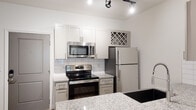 Located at 1500 Isner Ln in Garner, NC, this single-floor residence offers a thoughtfully designed layout with modern finishes throughout. The kitchen is a standout feature, equipped with a full suite of stainless steel appliances — including an electric range, over-the-range microwave, refrigerator, and dishwasher — complemented by granite countertops, white subway tile backsplash, white shaker-style cabinetry, and a deep farmhouse-style sink with a coil-spring faucet. A decorative wine rack accent adds charm. Track lighting illuminates both the kitchen and the open living area, which features light wood-look flooring, built-in shelving, and a frosted glass interior door that adds a contemporary touch. Bedrooms feature plush carpeting and large windows that invite abundant natural light with views of the surrounding area. Each bedroom is accompanied by a spacious walk-in closet outfitted with extensive wire shelving systems for organized storage. The bathrooms are finished in crisp white tile, with one offering a soaking tub/shower combination and another featuring a walk-in shower, both with brushed nickel fixtures and built-in corner shelving. The entry foyer provides a clean transition into the living spaces, with durable wood-look flooring continuing throughout the common areas.
