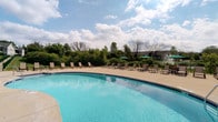 This single-story community clubhouse is thoughtfully designed to serve as a central hub for socializing and relaxation. The exterior showcases attractive stone masonry accents, green architectural columns, and an arched entryway, complemented by a pergola and a generously sized patio furnished with comfortable wicker seating — ideal for outdoor gatherings.

Inside, the main gathering room impresses with soaring vaulted ceilings and abundant natural light streaming through large windows. The open layout accommodates multiple dining tables and connects seamlessly to a full kitchen equipped with a refrigerator, microwave, bar-height seating, and ample counter space — well-suited for community events and catered functions.

Adjacent to the main room, a bright and inviting lounge area features a dramatic floor-to-ceiling stacked stone fireplace that serves as a striking focal point. Leather armchairs and expansive windows offering views of the surrounding greenery create a warm, comfortable retreat.

Outdoors, the spacious swimming pool area features a gently curved pool with crystal-clear water, surrounded by a broad concrete deck lined with lounge chairs and shaded umbrella tables. Lush landscaping and black iron fencing frame the space, providing both privacy and a park-like atmosphere.
