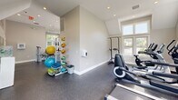 Located at Deer Creek in Plainsboro, NJ, this fitness center offers a thoughtfully designed, single-floor exercise facility that provides residents with a comprehensive workout environment. The exercise room is the centerpiece of the space, characterized by soaring vaulted ceilings and an abundance of natural light streaming through multiple large windows on all sides. Glass entry doors open directly to the outdoors, further enhancing the bright, airy atmosphere. Durable rubber flooring in a dark speckled finish covers the entire workout area, providing both safety and comfort underfoot. The facility is well-equipped with a Cybex functional trainer, a power tower, multiple cardio machines including a stationary bike and treadmills, an adjustable weight bench, a dumbbell rack, and a full medicine ball storage station. A large wall-mounted mirror along one side of the room reflects the space and aids in exercise form. Recessed lighting supplements natural light throughout. A dedicated bathroom completes the facility, finished with white subway tile wainscoting, brown floor tile, stainless steel grab bars for accessibility, and a frosted privacy window. The overall ambiance is clean, modern, and welcoming, making this fitness center a practical and well-appointed amenity.
