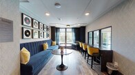 Located at 360 Creamery Way in Exton, Pennsylvania, this single-floor lounge space offers a sophisticated and music-inspired atmosphere that blends retro charm with contemporary design. The room is anchored by a long, channel-tufted navy blue velvet banquette, accented with gold throw pillows, positioned beneath a striking gallery wall of framed vinyl records. Complementing this seating area is a sleek marble-top tulip-style table, adding a refined touch to the lounge's aesthetic.

Along the opposite wall, a built-in countertop bar-height workspace is lined with stylish gold and charcoal upholstered chairs, providing a functional yet elegant area for work or socializing. Large windows dressed with teal sheer curtains flood the space with natural light and offer views of the surrounding greenery, enhancing the open and airy feel of the room.

The flooring transitions between light hardwood in the main lounge area and rich, warm-toned hardwood near the entryway, where an additional gallery wall showcases framed classic album covers—a nod to iconic music history. A coffered ceiling detail with a modern ceiling fan adds architectural interest overhead. The overall ambiance is inviting and curated, balancing bold design elements with comfortable, functional furnishings throughout.