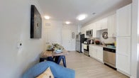 This single-floor residence offers a thoughtfully designed open-concept layout that seamlessly connects the living, dining, and kitchen areas into one cohesive and functional space. Light wood-tone flooring runs throughout, lending a clean and contemporary feel to the interior.

The kitchen is a standout feature, outfitted with crisp white shaker-style cabinetry, dark stone countertops, and a full suite of stainless steel appliances, including a refrigerator, range, and microwave. The cabinetry provides generous storage, while the layout allows for efficient use of the space.

Adjacent to the kitchen, the dining area accommodates a rustic wood dining table with bench seating, creating an inviting spot for meals. A statement wall-mounted piece of artwork adds a decorative focal point to the shared living and dining zone.

The living area flows naturally from the dining space, with comfortable seating and warm accent pillows that add a welcoming touch. Recessed and flush-mount ceiling lighting ensures the entire open-plan area is well illuminated throughout the day.

The entryway, visible from the main living space, features clean white paneled doors and maintains the consistent, modern aesthetic found throughout the residence. Overall, the space strikes a balance between functionality and modern style.