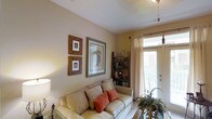 This single-floor space offers a warm and inviting atmosphere, thoughtfully designed for comfortable everyday living. The living area serves as the heart of the space, characterized by neutral-toned walls that create a calm, welcoming ambiance. Natural light streams in through a windowed door dressed with sheer white curtains and slatted blinds, brightening the room and lending an airy quality to the interior. The living area is furnished with a cushioned sofa accented by rust-colored throw pillows, complementing the earthy, muted color palette of the space. A collection of framed wall art in varying sizes and styles adds personality and visual interest to the walls, while a freestanding wooden bookshelf in the corner provides both storage and a decorative focal point, displaying books and small decorative objects. Overhead, a ceiling fan ensures year-round comfort. A floor lamp and potted greenery contribute to the cozy, lived-in character of the room. The overall design aesthetic blends casual comfort with thoughtful decorative details, making this a welcoming and functional single-floor living space.