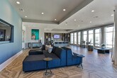 Located at 6000 Summit Place in Alpharetta, Georgia, this single-floor amenity space offers a sophisticated and contemporary design that blends comfort with modern aesthetics. The standout architectural feature throughout is the striking chevron-patterned hardwood flooring, which anchors the entire space with warmth and visual interest.

The lounge area is anchored by a bold blue accent wall housing a wall-mounted flat-screen television, complemented by a deep navy sectional sofa and minimalist side tables. Curated artwork, including a vibrant large-scale painting, adds a gallery-like character to the space. Glass double doors provide connectivity to adjacent areas and allow natural light to flow freely throughout.

Transitioning into the kitchen and gathering zone, a large white island with seating for multiple guests serves as a central hub. Dark open shelving units frame the area, displaying decorative accents and providing practical storage. Pendant lighting suspended from a tray ceiling highlights the island, while floor-to-ceiling windows along the perimeter flood the space with abundant natural light and offer views of the surrounding Alpharetta streetscape.

The open-plan layout across the entire space encourages flexible use, from casual socializing to organized events. Recessed lighting throughout ensures consistent illumination, and the neutral wall palette contrasts elegantly with the rich tones of the flooring and furnishings.