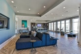 Located at 6000 Summit Place in Alpharetta, Georgia, this single-floor amenity space offers a sophisticated and contemporary design that blends comfort with modern aesthetics. The standout architectural feature throughout is the striking chevron-patterned hardwood flooring, which anchors the entire space with warmth and visual interest.

The lounge area is anchored by a bold blue accent wall housing a wall-mounted flat-screen television, complemented by a deep navy sectional sofa and minimalist side tables. Curated artwork, including a vibrant large-scale painting, adds a gallery-like character to the space. Glass double doors provide connectivity to adjacent areas and allow natural light to flow freely throughout.

Transitioning into the kitchen and gathering zone, a large white island with seating for multiple guests serves as a central hub. Dark open shelving units frame the area, displaying decorative accents and providing practical storage. Pendant lighting suspended from a tray ceiling highlights the island, while floor-to-ceiling windows along the perimeter flood the space with abundant natural light and offer views of the surrounding Alpharetta streetscape.

The open-plan layout across the entire space encourages flexible use, from casual socializing to organized events. Recessed lighting throughout ensures consistent illumination, and the neutral wall palette contrasts elegantly with the rich tones of the flooring and furnishings.