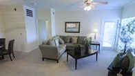 This single-floor residence offers a thoughtfully designed layout that balances comfort and functionality. The living area serves as the heart of the space, featuring soft neutral-toned carpet, crisp white walls accented with elegant crown molding, and a ceiling fan that adds both style and practical comfort. Natural light filters in through a door to the exterior, brightening the room and creating an inviting atmosphere. A framed wall mirror reflects a glimpse of the kitchen beyond, hinting at an open and connected floor plan. The living area is generously proportioned, providing ample room for a large sectional sofa, accent chair, and a substantial coffee table — all centered around a cohesive, neutral color palette. A side table with a classic lamp adds a warm, finished touch to the seating arrangement. Visible through the hallway, additional rooms extend the living space further, suggesting a practical and well-organized layout. The kitchen, partially visible in the mirror's reflection, appears bright and equipped with modern appliances. Overall, the space presents a clean, comfortable, and move-in-ready environment with classic architectural details that lend a timeless, welcoming character throughout.