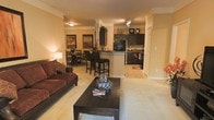 This single-floor residence offers an open-concept design that brings together the living, dining, and kitchen spaces in one cohesive and inviting layout. The living area is anchored by plush, comfortable seating and a dark wood coffee table, all set atop soft wall-to-wall carpeting that adds warmth underfoot. Crown molding along the ceilings lends a refined architectural touch throughout the space.

The kitchen is a standout feature, equipped with dark cabinetry, a black refrigerator, and a raised breakfast bar with seating for multiple guests — perfect for casual dining or entertaining. The kitchen flooring transitions to tile, providing a clean contrast to the carpeted living and dining areas. Track lighting overhead ensures the kitchen is well-lit and functional.

Adjacent to the kitchen, a dedicated dining area accommodates a full dining table and chairs, ideal for more formal meals. Warm, golden-toned walls carry throughout the open layout, creating a cohesive and cozy ambiance. Decorative accents, including framed artwork and greenery above the cabinetry, add personality and character to the space. Overall, the home balances everyday functionality with comfortable, inviting style.