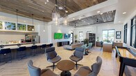 This single-floor community clubhouse delivers a thoughtfully designed, open-concept layout that seamlessly blends social and leisure functions. The space is anchored by a striking interior aesthetic that pairs crisp white walls with warm, reclaimed wood ceiling panels, creating a balanced contrast between modern and rustic elements. Light oak herringbone-patterned flooring runs throughout, adding visual texture and cohesion across each zone.

The kitchen and bar area serves as a central gathering point, featuring a large white quartz island with navy blue bar stools, complemented by stainless steel appliances, a tile backsplash, and a dedicated beverage station. Vibrant botanical artwork displayed above the cabinetry adds a lively, curated touch. Pendant lighting illuminates the counter, enhancing the inviting atmosphere.

Adjacent lounge areas are furnished with an eclectic mix of swivel chairs in teal and gray tones, arranged around low tables to encourage conversation. Statement glass pendant fixtures hang above, while recessed lighting ensures the entire space is well-lit. A dedicated media lounge features dual wall-mounted flat-screen TVs with comfortable seating and ottoman poufs for a relaxed viewing experience.

Toward the rear, large black-framed windows flood the space with natural light and provide views to the exterior. A shuffleboard table adds a recreational focal point, making this clubhouse an ideal venue for both casual gatherings and organized social events.