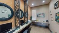 The Learning Jungle is a thoughtfully designed single-story study space that blends functionality with vibrant, nature-inspired décor. The room features warm terracotta and taupe walls adorned with framed wildlife and botanical artwork, including eye-catching leopard and tropical leaf prints that reinforce the jungle theme. Distinctive dark-framed lattice divider panels with a honeycomb pattern visually separate individual computer workstations, each equipped with a desktop computer, keyboard, and ergonomic seating. Striking round mirrors with textured dark frames are mounted above each station, adding depth and a stylish focal point to the workspace. A continuous built-in countertop runs along one wall, providing additional workspace that includes a shared printer and decorative accents like a teal grass plant arrangement. Natural light filters in through a window fitted with blinds, complementing the recessed ceiling lighting overhead. Light wood-tone flooring ties the space together, creating a clean, modern foundation. A small children's activity table with a wire bead maze is positioned near the entrance, making the space welcoming for all ages. A glass-paneled entry door offers a view to an outdoor area, further connecting the interior to a sense of the natural world.