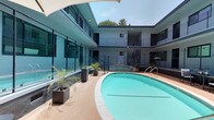 This contemporary property is centered around a stunning open-air courtyard that serves as its architectural focal point. The courtyard features a generously sized swimming pool with vivid turquoise water, framed by a clean, light-toned deck ideal for relaxation. Sleek lounge seating and candle holders are positioned poolside, offering a refined setting for leisure. Transparent glass pool fencing enhances both safety and sightlines throughout the space.

The surrounding structure rises to two stories, with each level connected by open staircases and finished with modern metal railings. Large floor-to-ceiling glass windows and sliding doors line the ground level, flooding interior spaces with natural light and maintaining a visual connection to the courtyard. The upper level features an open walkway with steel balustrades, providing an elevated view of the pool area below.

Tropical potted palms and decorative planters are strategically placed around the deck, adding a lush, resort-inspired quality to the space. The building's exterior is finished in a clean white and gray palette, complemented by dark-framed windows that lend a modern, polished aesthetic. Natural light pours into the courtyard from above, reinforcing the open and airy character of the entire property.