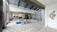 This single-floor lobby showcases a thoughtfully designed interior that blends coastal and contemporary aesthetics. The space is defined by its striking exposed dark wood beams set against a vivid blue ceiling, creating a dramatic architectural contrast with the otherwise light and airy surroundings. Large-format patterned tile flooring anchors the space, featuring a subtle swirling motif that adds visual texture underfoot.

The reception desk runs prominently along the back wall, finished with warm wood paneling and backlit displays, and equipped with multiple workstations to accommodate guests efficiently. Recessed lighting throughout ensures the space remains bright and inviting. A feature wall with alternating blue and natural wood horizontal panels adds a bold focal point near the entrance, complementing the overall coastal palette.

The seating area features a striped upholstered sofa in neutral tones paired with vibrant blue accent furniture and metallic side tables, striking a balance between comfort and style. Decorative sculptures and coastal artwork are tastefully placed throughout, reinforcing the design theme. The overall ambiance is open, polished, and welcoming, suited to a professional hospitality or commercial environment.