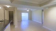 This single-story residence offers a clean, open floor plan designed for comfortable living. The main living area is a standout feature, characterized by a tray ceiling that adds architectural depth and dimension to the room. A ceiling fan is centrally mounted, providing both function and a finishing touch to the space. Large sliding glass doors dressed with vertical blinds allow abundant natural light to flood the interior, creating a bright and airy atmosphere throughout.

The flooring throughout the main living area consists of light-toned ceramic tile, which is both durable and easy to maintain. Neutral warm beige walls complement the tile and contribute to the overall sense of openness and cleanliness. Structural columns and half-walls subtly define the transition between living spaces while preserving the open feel of the layout.

Multiple doorways branch off from the main area, suggesting well-connected adjacent rooms that flow naturally from the central living space. The overall design is straightforward and functional, with architectural details like the tray ceiling elevating the interior beyond the purely utilitarian. The combination of natural light, open sightlines, and cohesive neutral finishes makes this a welcoming and versatile space suited for a variety of living arrangements.