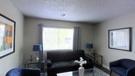 This single-floor space offers a thoughtfully arranged living area designed for both comfort and functionality. The living room serves as the heart of the space, centered around a sleek dark sofa complemented by matching accent tables and coordinating blue table lamps that add a touch of color and warmth. A large picture window dressed with gray grommet-top curtains and horizontal blinds allows generous natural light to filter in, creating a bright and inviting atmosphere with views of the surrounding greenery outside. The textured popcorn ceiling adds a classic architectural detail overhead, while neutral-toned walls provide a versatile backdrop that pairs well with the contemporary furnishings. A geometric-patterned area rug anchors the seating area, adding visual interest to the flooring beneath. A glass-topped coffee table and side tables lend a modern, airy feel to the room without overwhelming the space. The overall ambiance is clean, relaxed, and well-lit, making this a practical and comfortable living environment suitable for a variety of lifestyles.